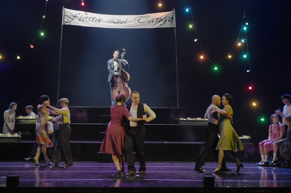 Max Dollinger als Augustin Magaldi in "Evita" mit Kontrabass auf einer Bühne, davor tanzende Tango-Paare, Bühnenbild