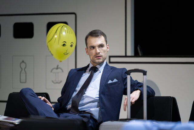 Mann in feinem Anzug sitzt auf einem Stuhl im Wartesaal, neben ihm ein Koffer und ein Luftballon mit bemalten Gesicht